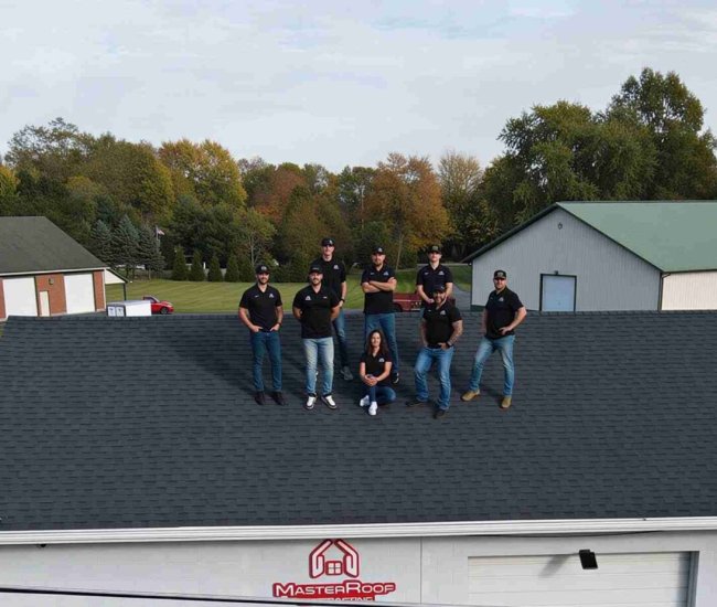 Nine workers stand on a dark shingle roof of a Master Roof building with a suburban complex in the background on a clear day.