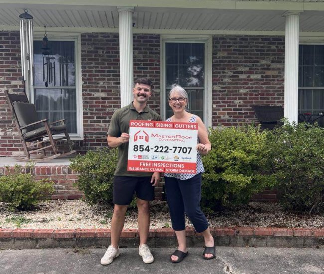 Smiling man and woman stand on a sidewalk in front of a brick house, holding a roofing and gutters advertisement sign.