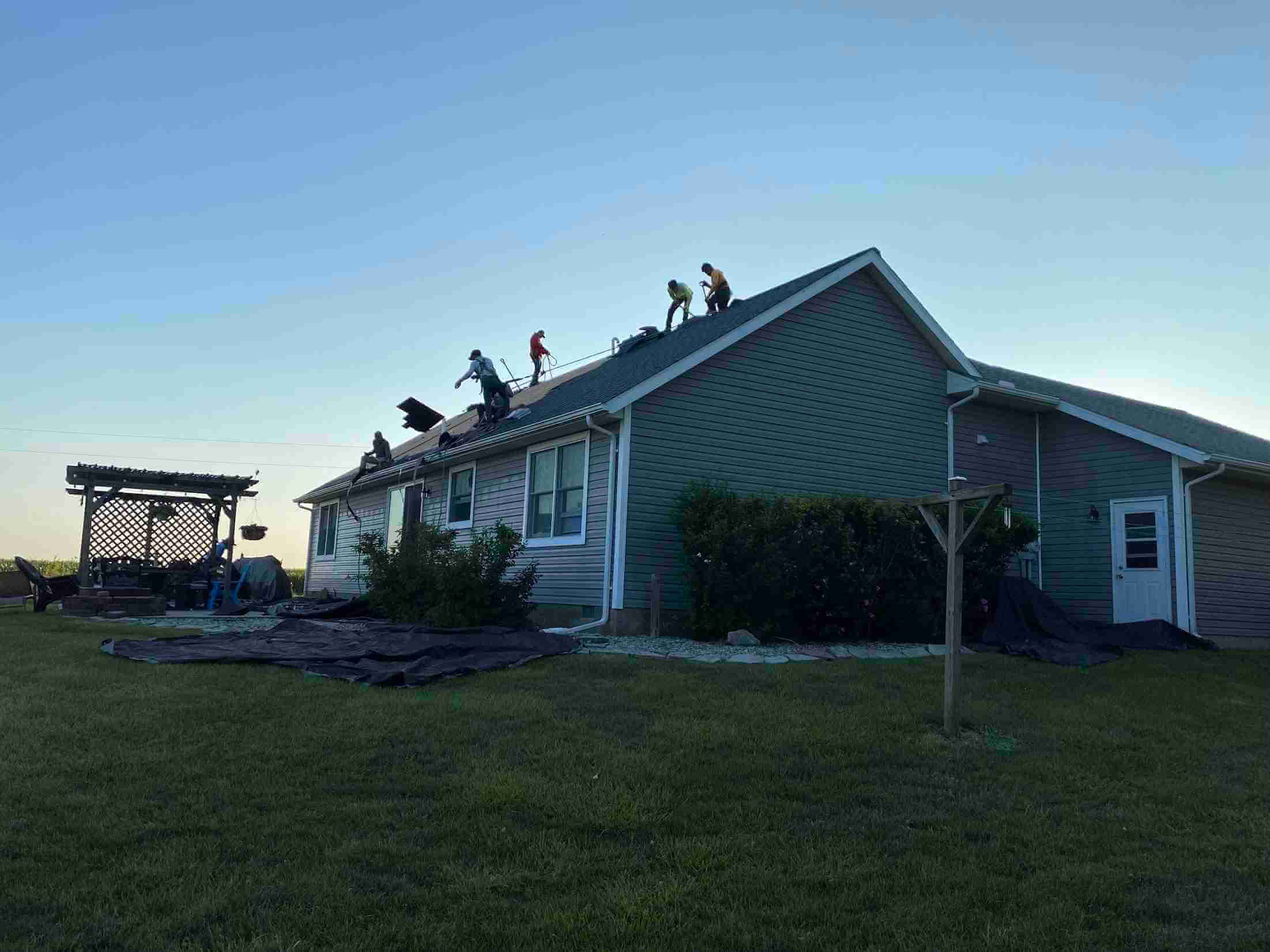 Roofers working on the sloped roof of a two-story house during daylight, with ladders and safety gear visible on the roof edge