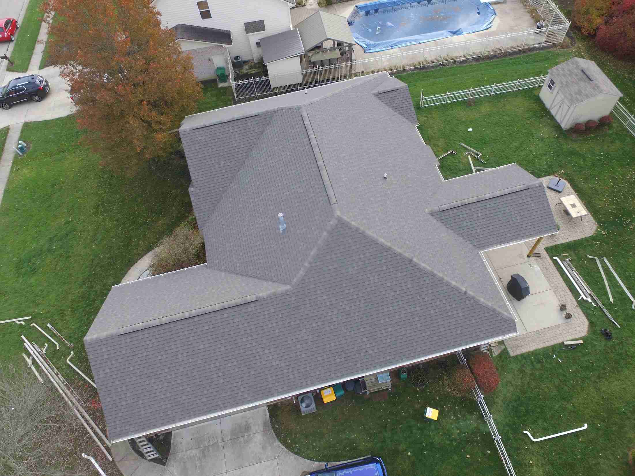 Aerial view of a house with a large gray roof, fenced yard, a covered pool, trees, and nearby parked cars.
