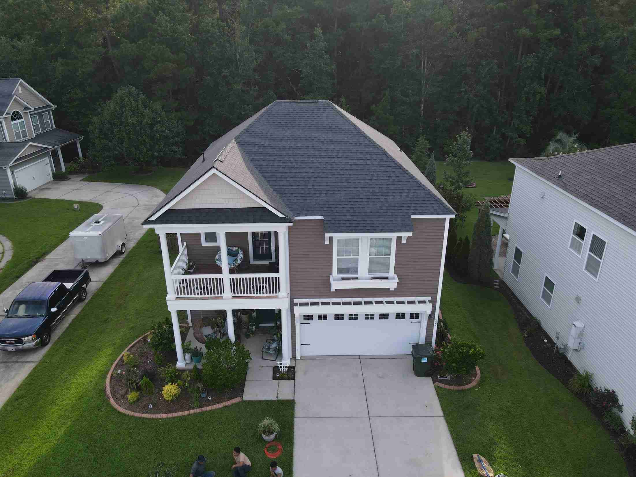 Aerial view of a two-story suburban home with a gray roof, front porch, and attached garage, surrounded by a green lawn and neighboring houses.