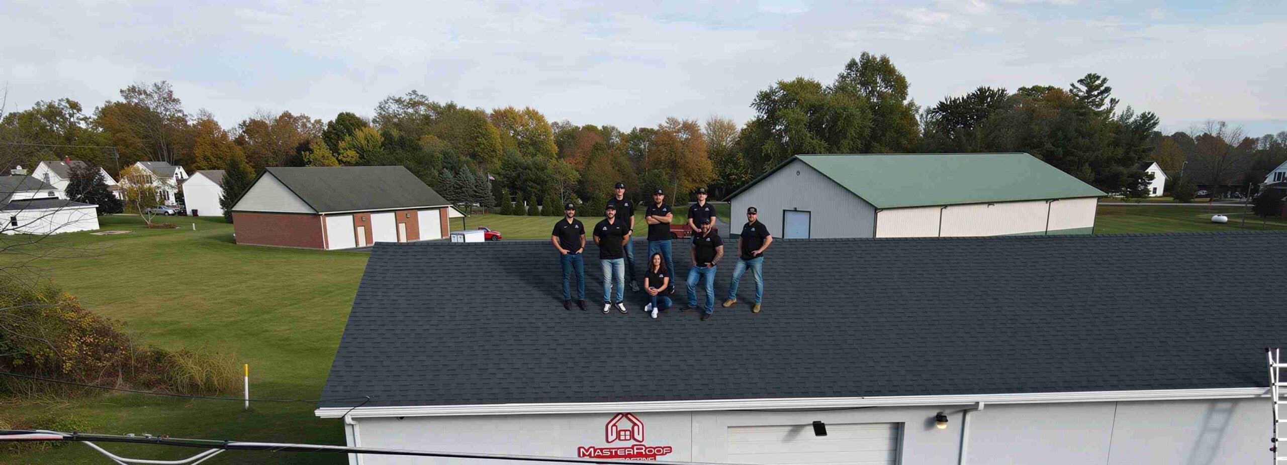 Nine workers stand on a dark shingle roof of a Master Roof building with a suburban complex in the background on a clear day.