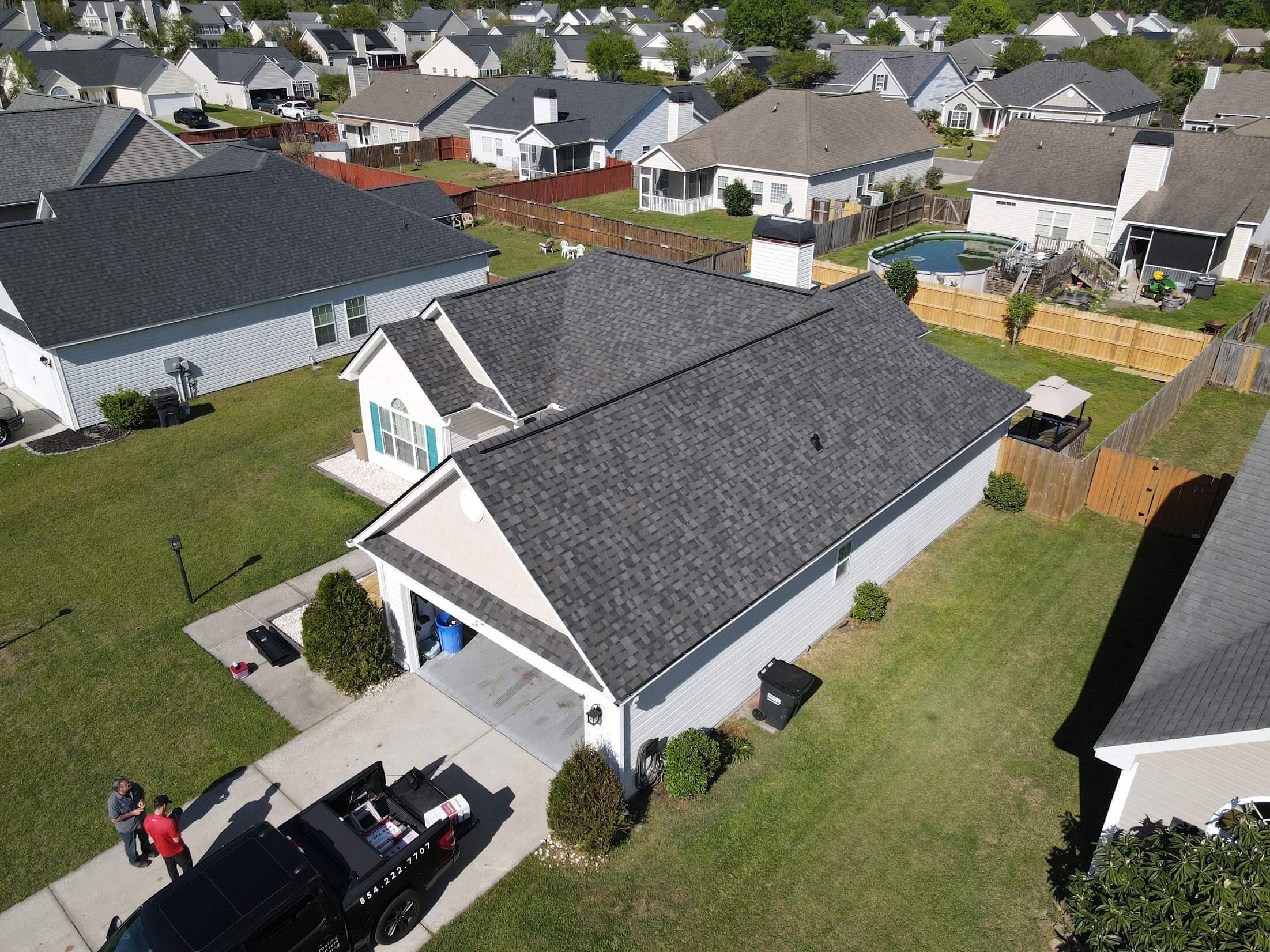 Aerial view of a suburban neighborhood with gray-shingled roofs, green lawns, and fenced backyards including a round pool in one yard setting the scene for a residential area