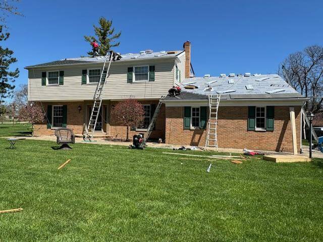Two-story brick house with workers on the roof and ladders, during roof replacement on a sunny day.
