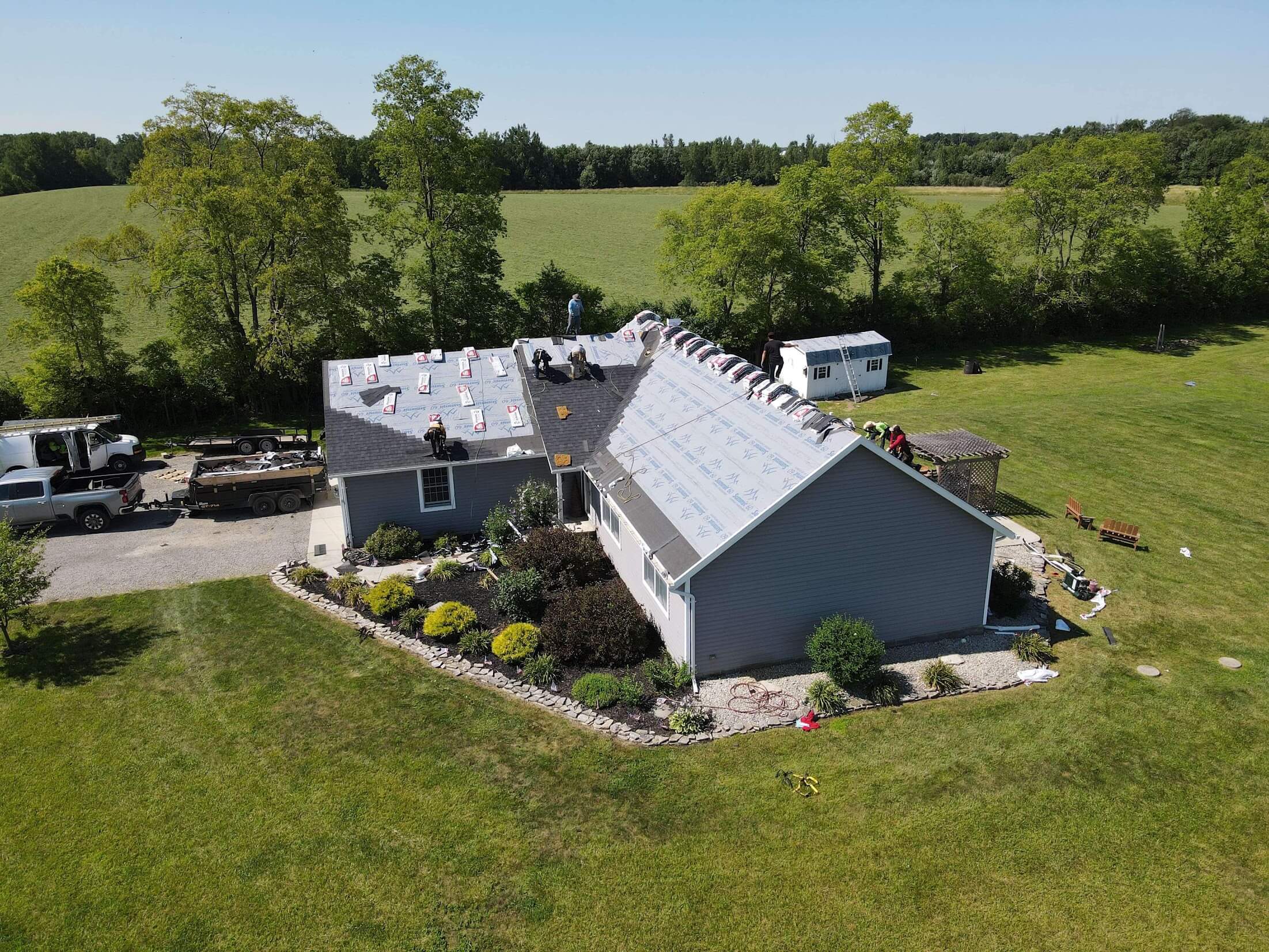 Aerial view of a house undergoing roof replacement, with workers on the roof and construction equipment and trucks in the driveway, set against a large grassy yard and trees in the background.