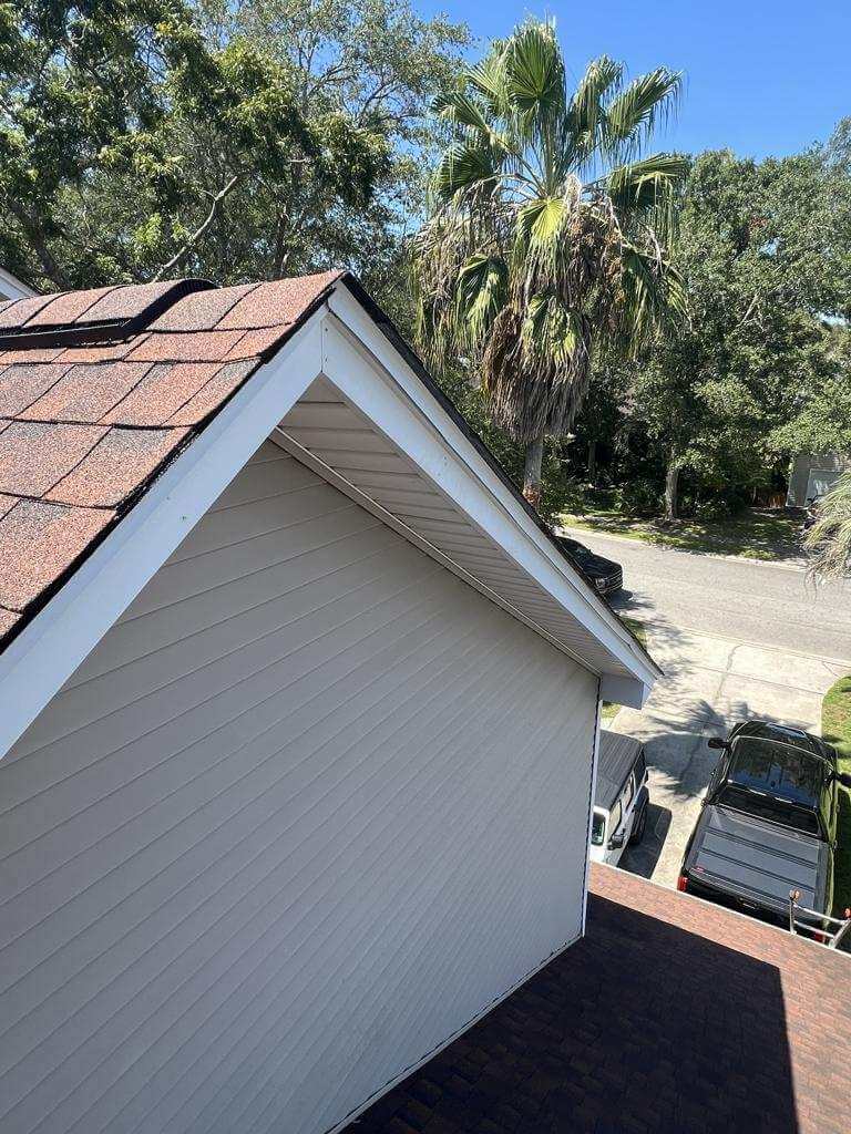 Aerial view of a white house wall with brown shingled roof, palm trees in the yard, and parked cars along a sunny street nearby.