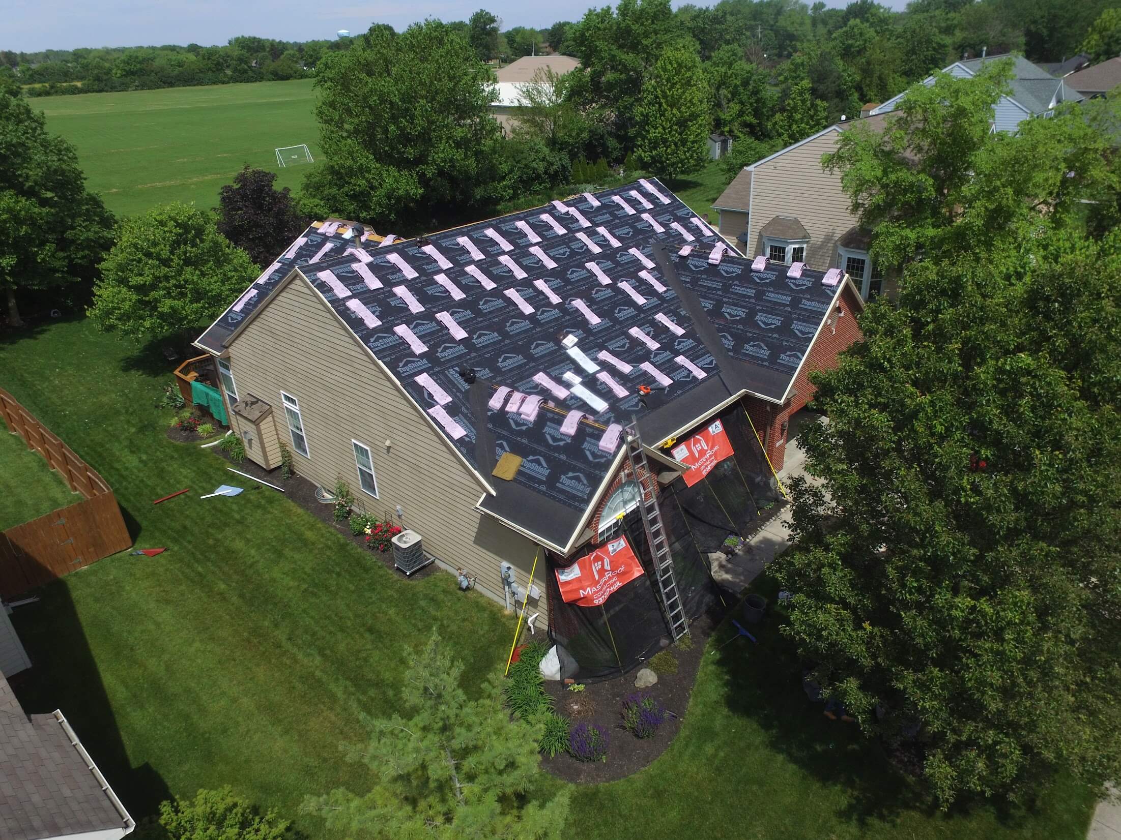 Aerial view of a house under roof construction with underlayment and pink insulation strips, ladders, and construction materials in the yard.