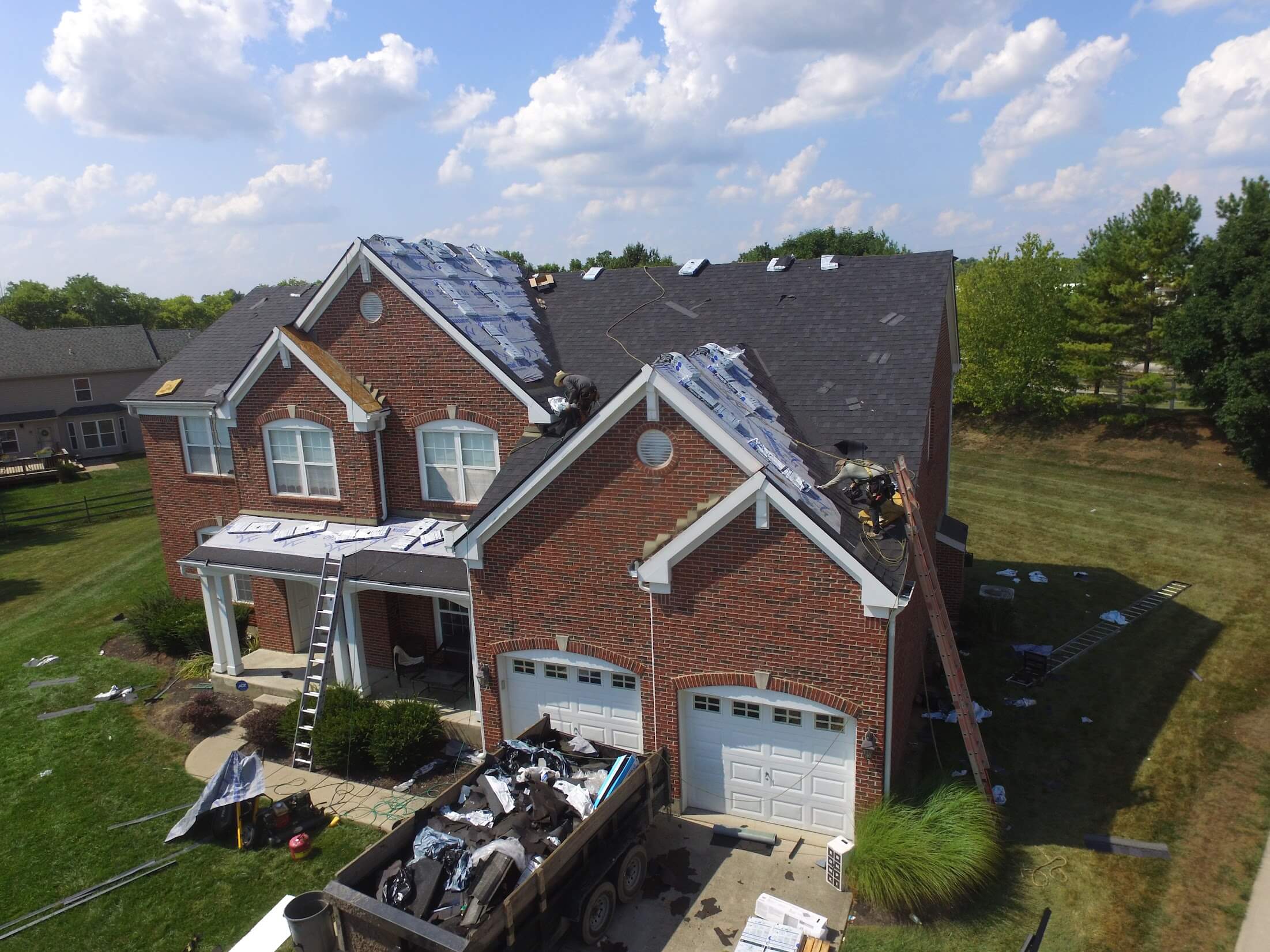 House with workers on the roof replacing shingles; a dump trailer and tools in the yard under a blue sky with clouds.