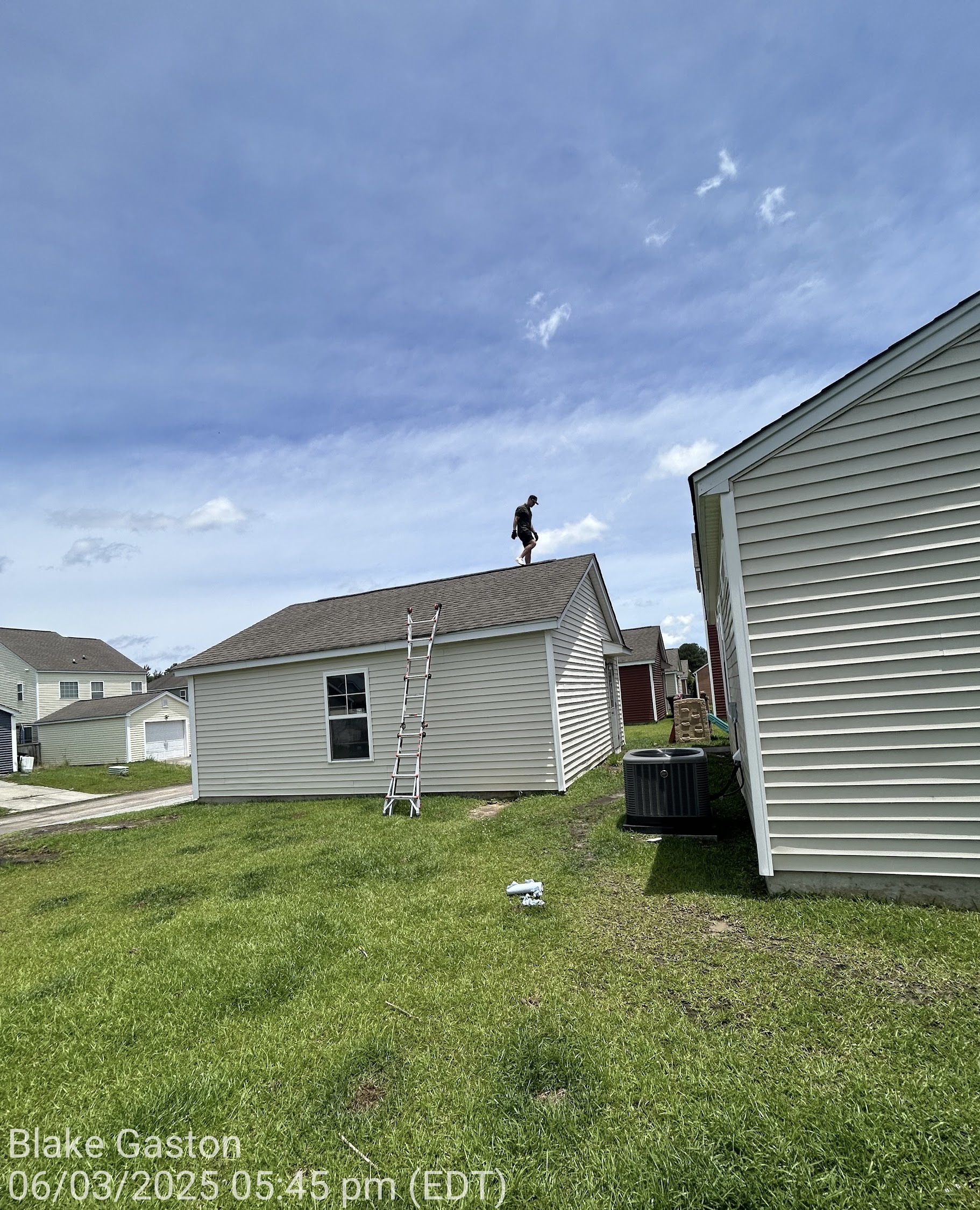 Man standing on the peak of a suburban house roof beside a ladder on a sunny day, with blue sky overhead and neighboring houses nearby.