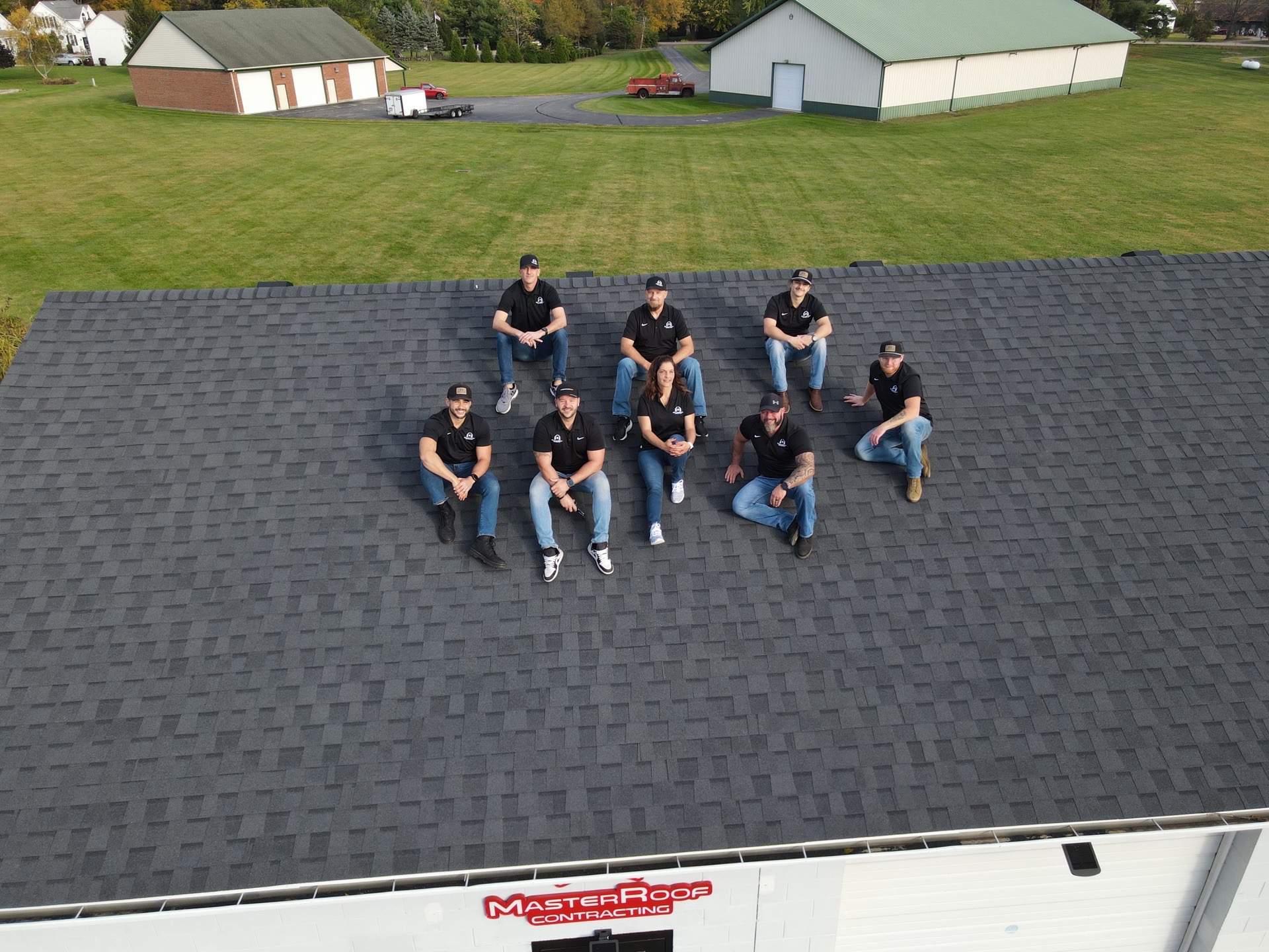 Aerial view of nine roofing crew members in black shirts sitting on a dark gray shingled roof, with a MasterRoof Contracting edge sign visible at bottom edge.