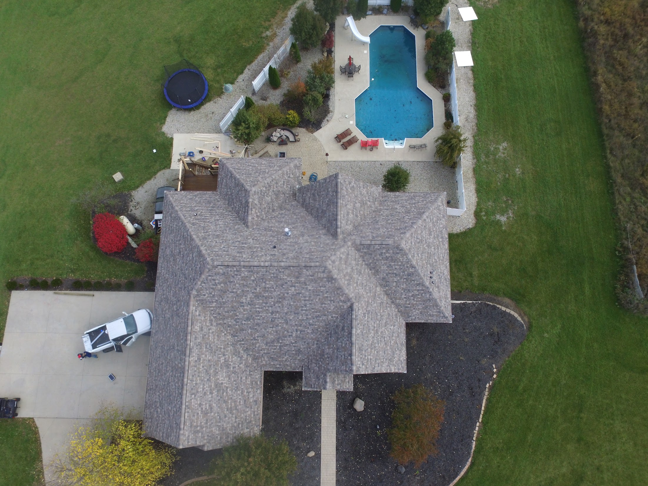 Aerial view of a large house with a blue swimming pool, surrounding patio, and green lawn. A trampoline and landscaping are visible on the left side.