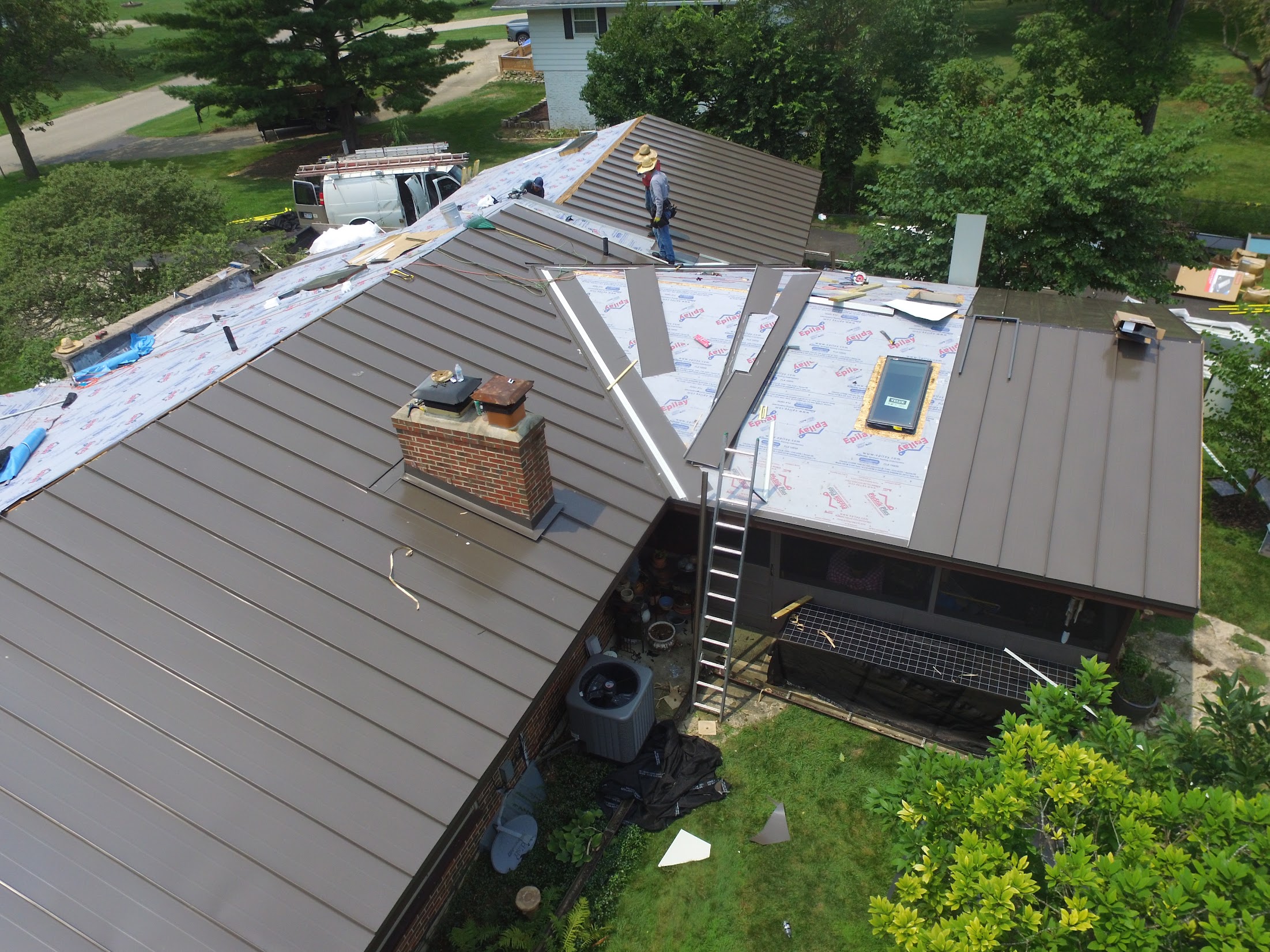 Construction workers installing metal roofing panels on a sloped roof; ladder and tools scattered, white van parked nearby in a yard.