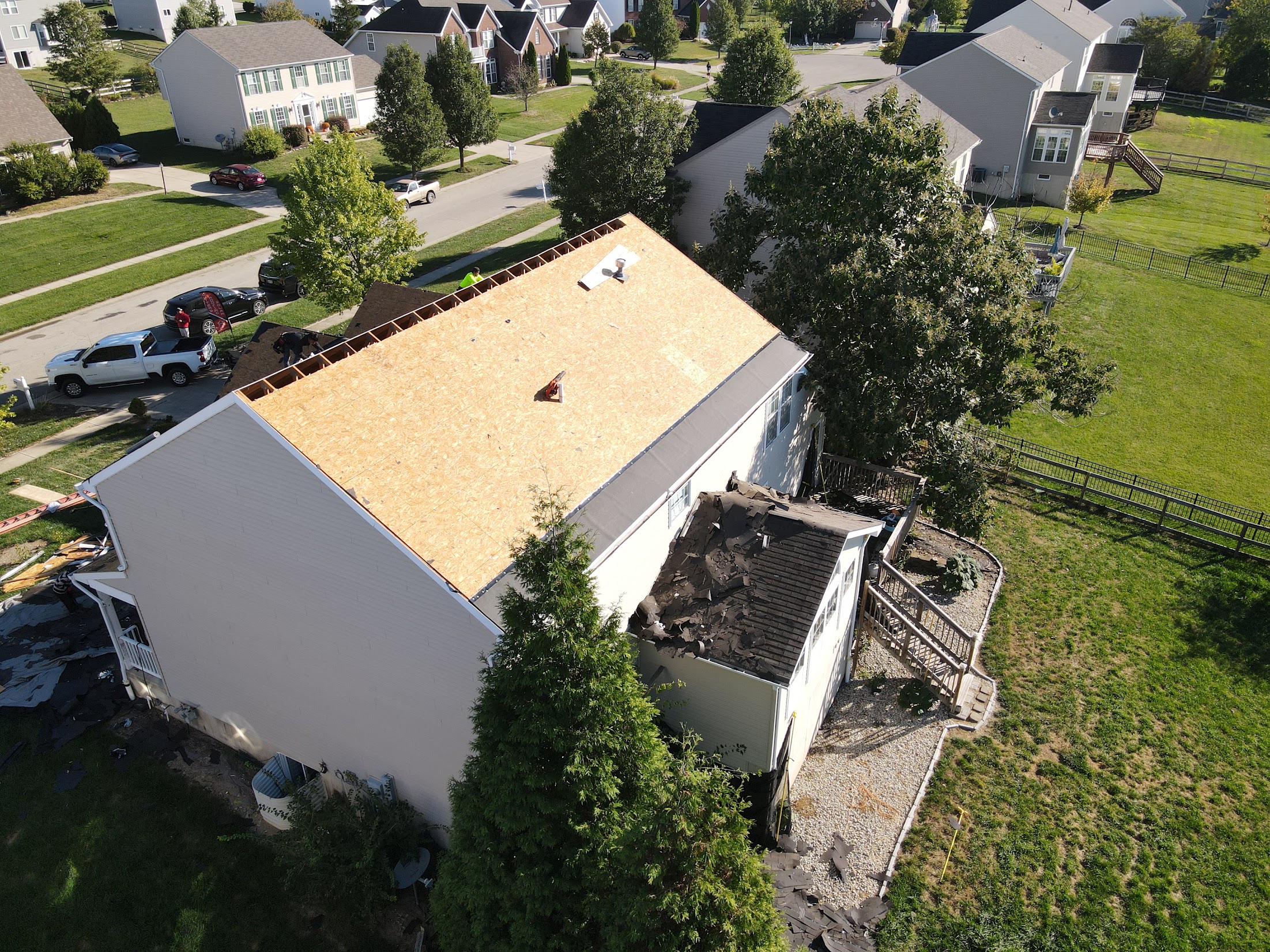 Aerial view of a two-story house under construction with a plywood roof and workers on the roof, surrounded by a suburban neighborhood.